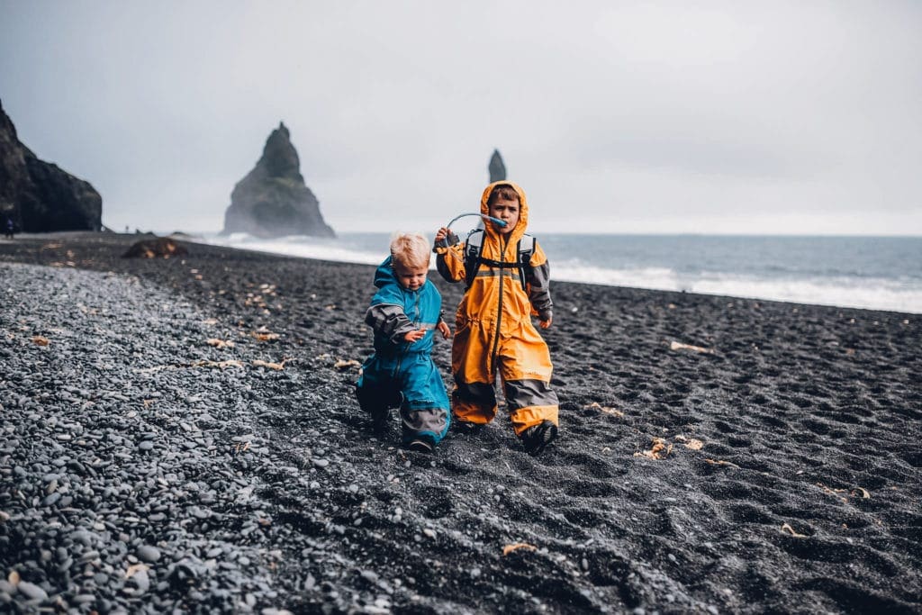 Two boys, one in yellow and one in blue, walk across a black beac in Iceland.