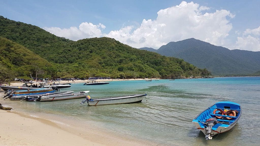 A boat rests along the shore at Tayrona Park in Colombia, with lush mountains in the distance.