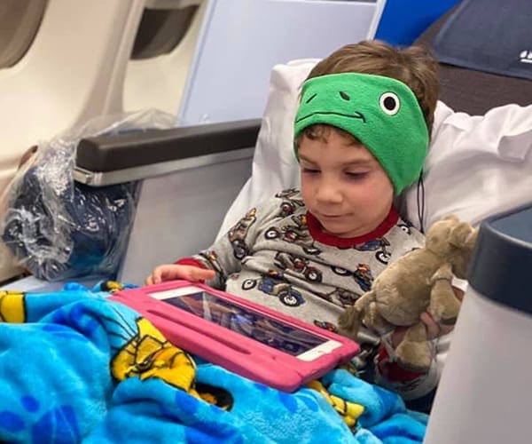 Child on airplane seat with blue blanket, stuffed animal and iPad in pink case with green headphones