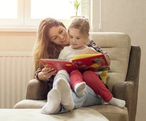 A mom and her little girl read a book together on a comfy chair.