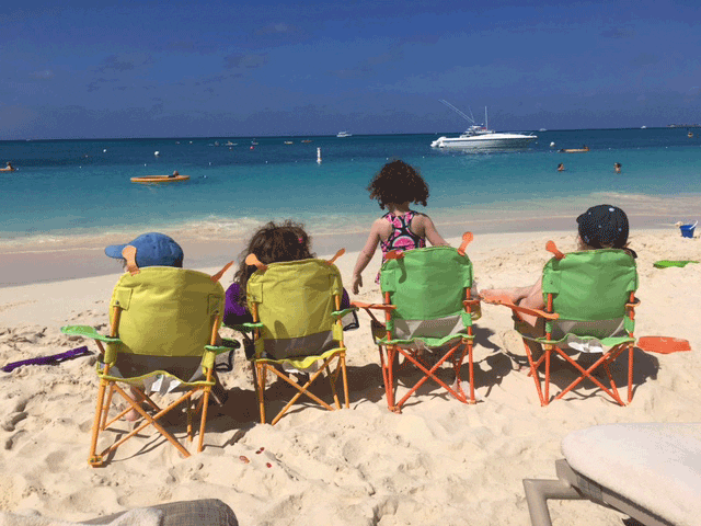 Four kids sit in a row in small beach chairs by water in Grand Cayman.