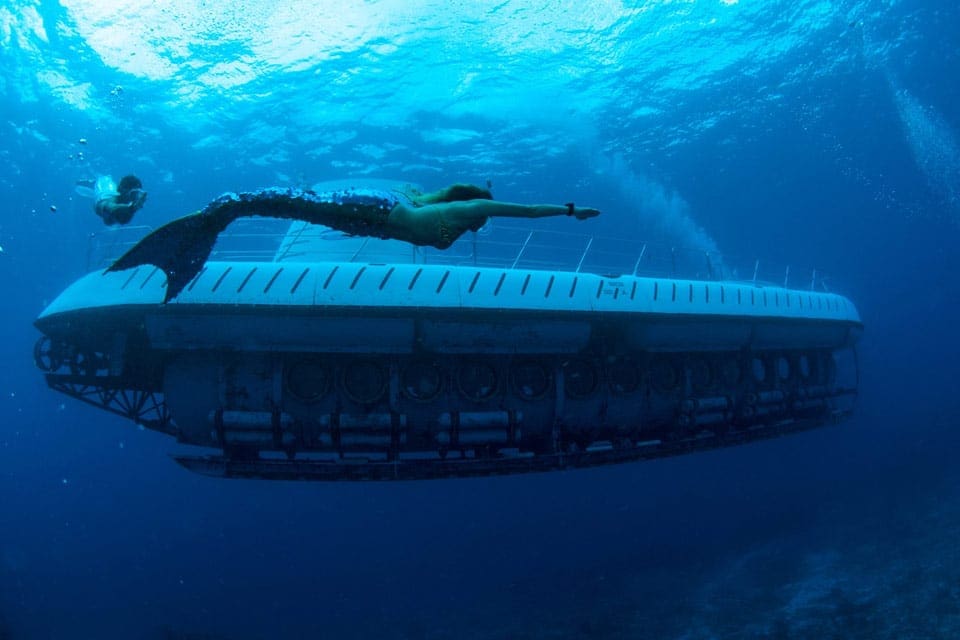 A mermaid swims by a submarine in a deep blue sea at Atlantis Submarine.