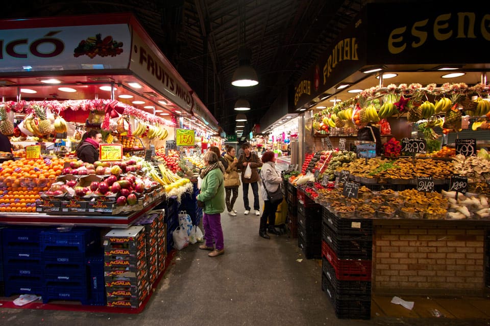 Inside the Boqueria Market, with stalls on either side of a sidewalk. In the distance, several shoppers look at produce and speak with vendors.