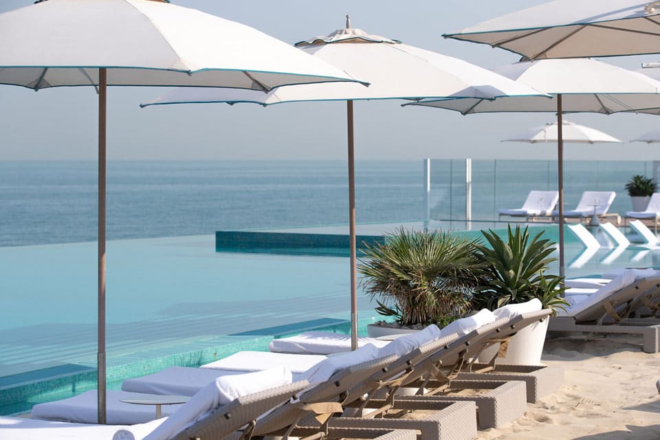 Pristine white oceanside loungers await guests under white umbrellas, while lined along the beachfront of Burj Al Arab, one of the best family hotels in Dubai.