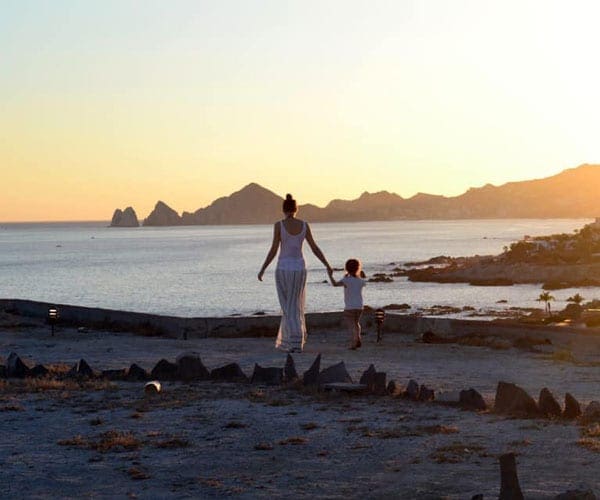 Mom wearing a beautiful while dress walking with a little boy holding hands with beach and sunset in the background