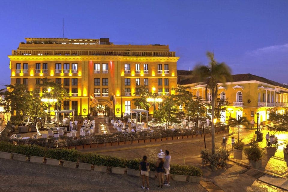 Three people standing on the street look toward the well-lit entrance to the Hotel Charleston Santa Teresa at night.