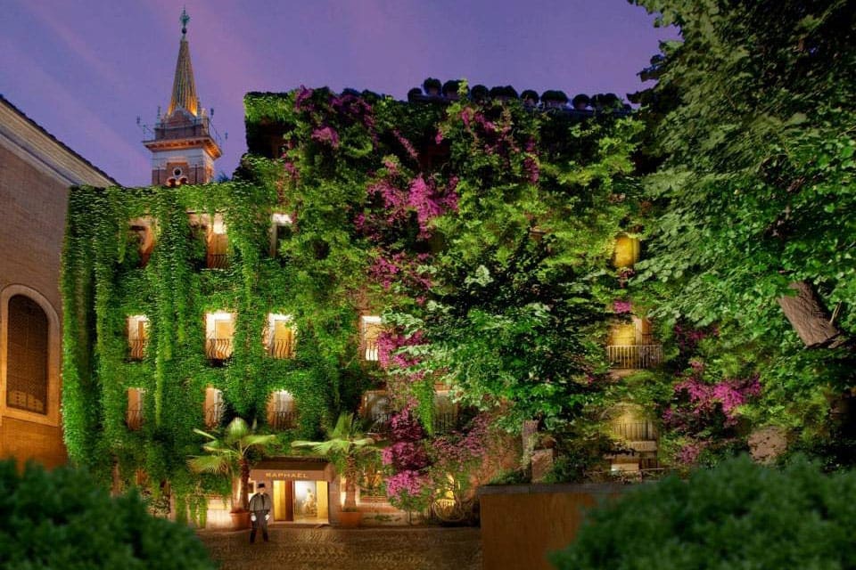 A staff member stands outside the door to J.K Place Roma, with vines hanging on the sides of the building, at dusk.