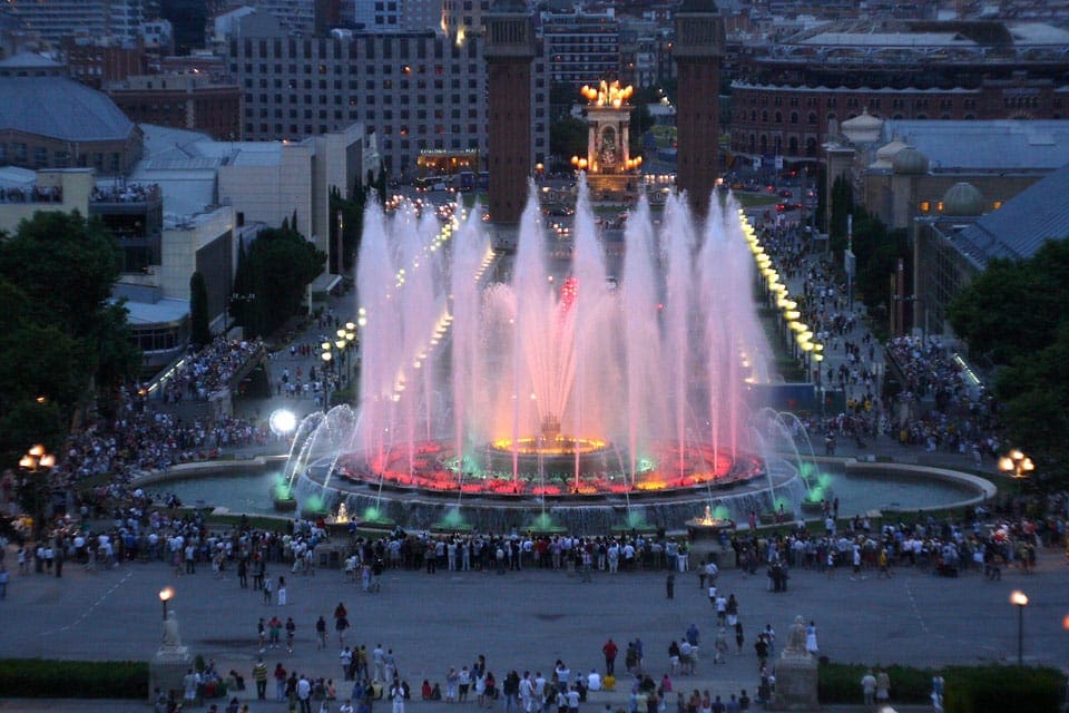 A colorful light display brightens the Magic Fountain of Montjuic, while a plaza of onlookers enjoy the show.