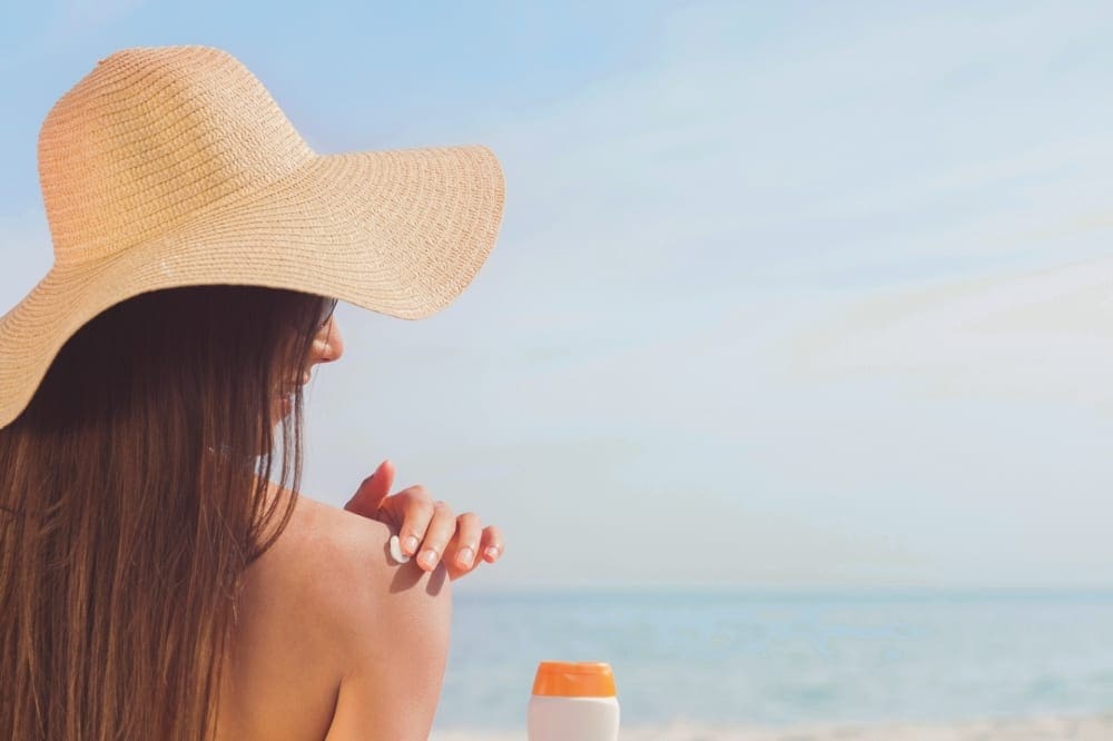 A woman applies sunscreen to her shoulder, while enjoying a day at the beach, sunscreen is a must on a beach packing list for families.