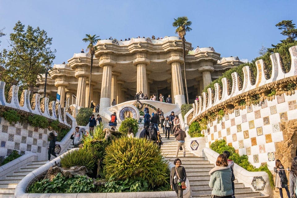 The outdoor entrance to Parc Guell, featuring its iconic stairs, Gaudi art, and palms.