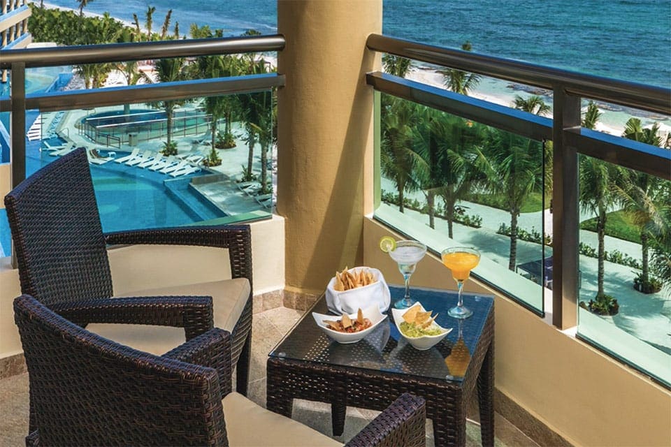 An inviting table holds two tropical drinks and snacks with a view of the pool and the ocean at Generations Riviera Maya, one of the best resorts in Mexico for families.