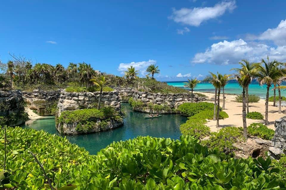 An aerial view of the sunny grounds of Hotel Xcaret, featuring beach access and a large pool flanked by swaying pines at one of the best all-inclusive resorts Playa del Carmen for families.