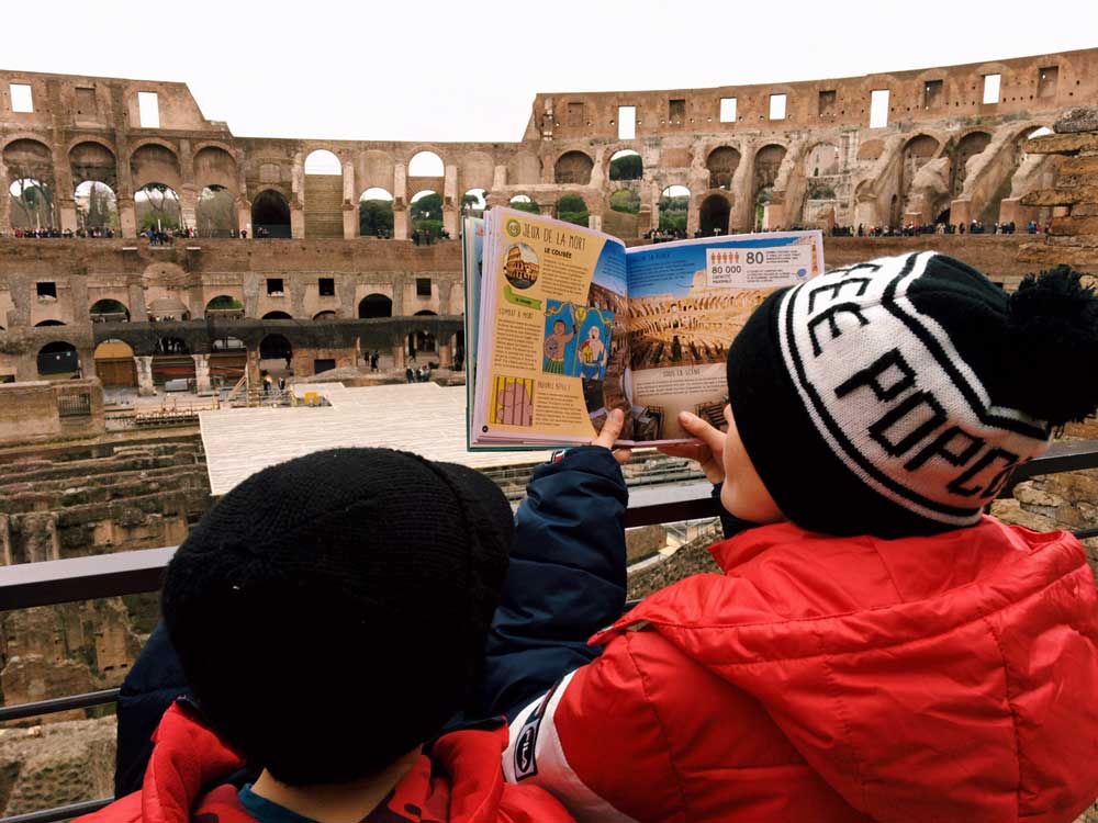 Two boys in looking at a book about the Colosseum, while standing within its grounds.