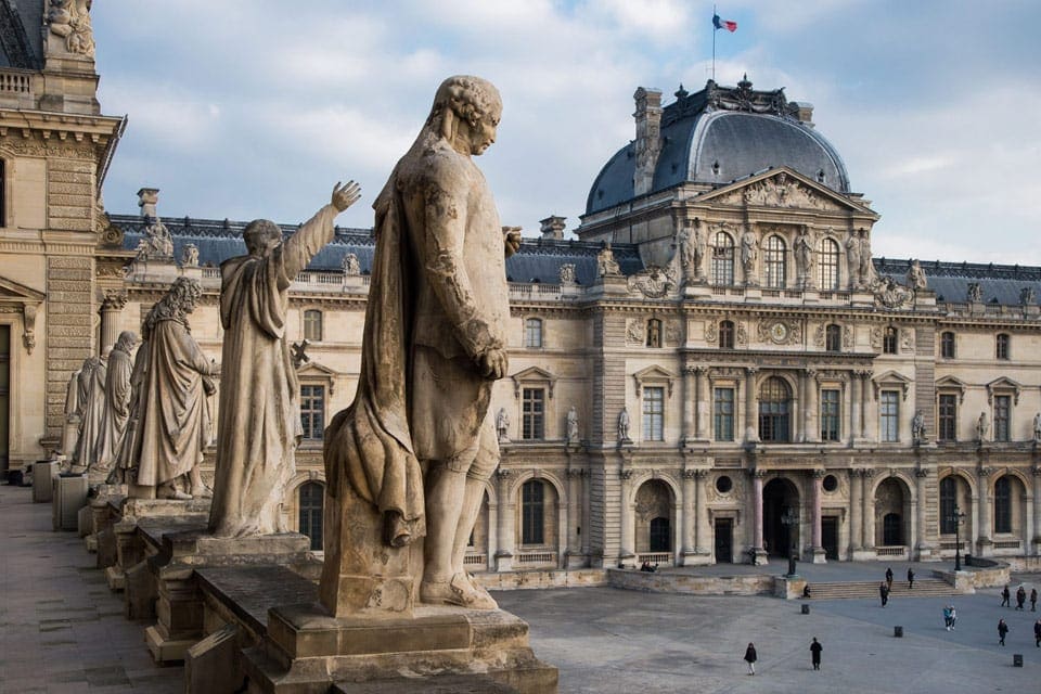 Statues atop the Louvre Museum looking down onto a courtyard in Paris.