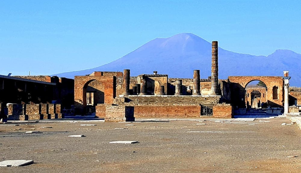 A view of ruins at Pompeii, with Mount Vesuvius looming in the background. Mount Vesuvius is one of the stops on our virtual vacation to Naples and Pompeii.