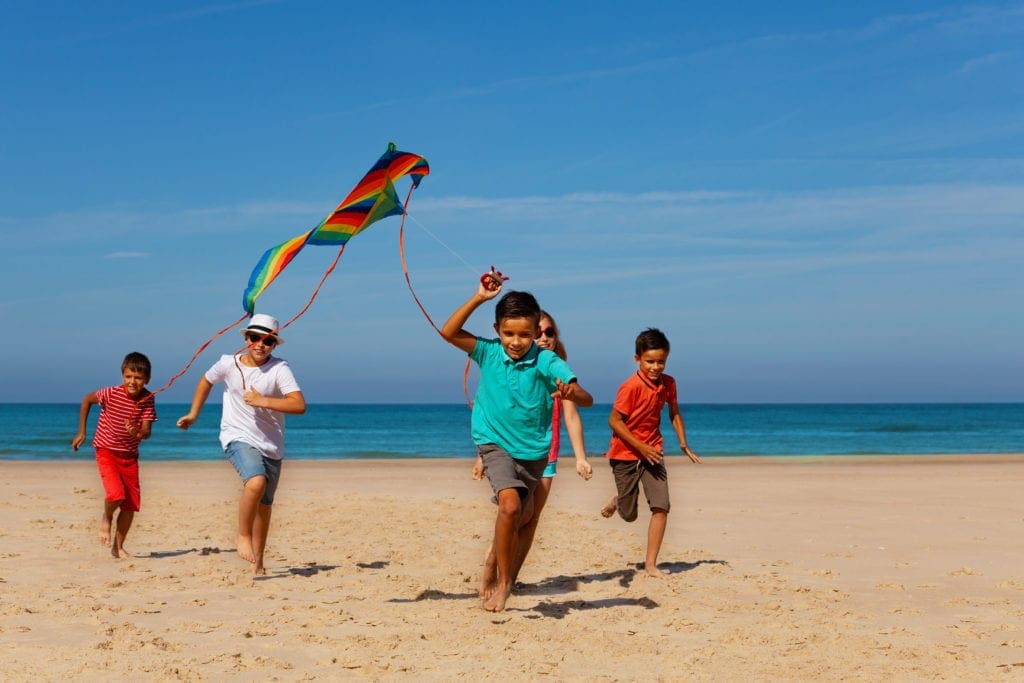 Group of happy children boys and girls run on the sand beach holding colorful flying kite with sea on background