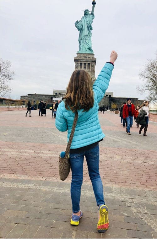 A young girl stands with her fist in the air facing the Stature of Liberty in the background, one of the best New York City outdoor activities for kids.