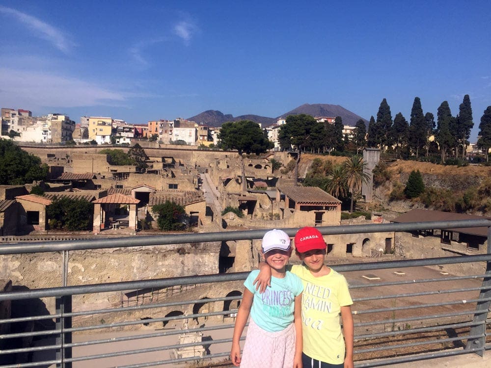 Boy and a girl stand together in Pompeii. Ruins dot the background while Mount Vesuvius looms in the distance. Pompeii is a must see on our virtual vacation to Naples and Pompeii.