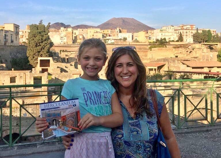 Mother and daughter stand smiling showing off their Herculaneum book. The ruins of Pompeii dot the background.