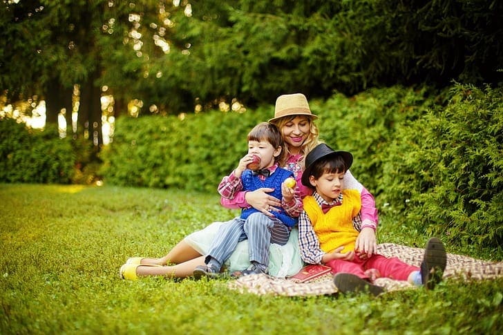 A mom sits with two kids on her lap in a lush park.