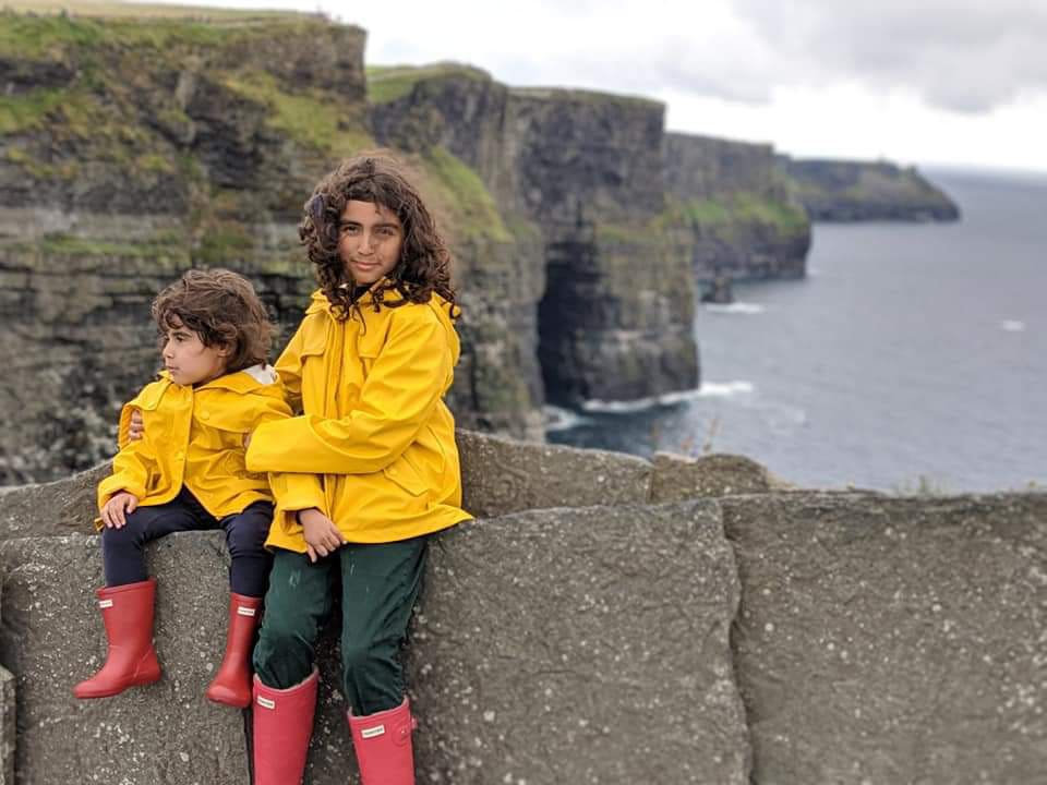 Two girls sitting together on cliff in Ireland, with a sweeping view of the cliffs and ocean behind them, while traveling on a family vacation.