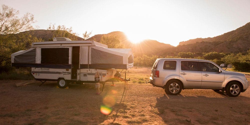 Pop-up camper being towed by a large vehicle sits as a desert campsite at sunset.