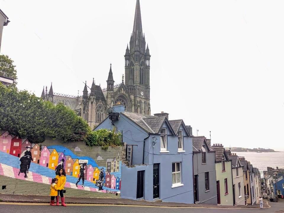 Two kids in matching yellow raincoats stand in front of a colorful mural on a street in Ireland.