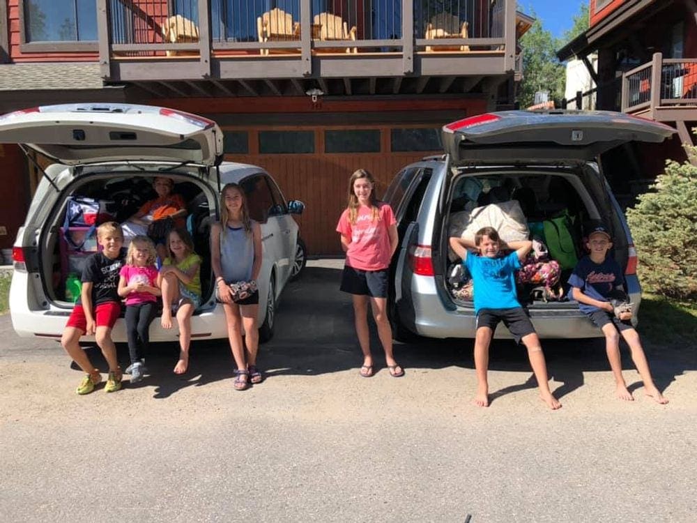 Seven kids stand in front of 2 packed vans in preparation of a road trip. Servicing your car before a trip is one of our tips for long car rides with toddlers