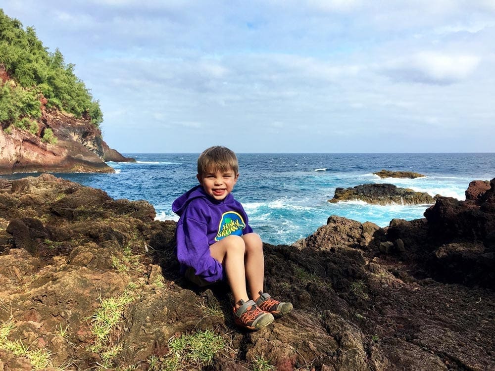 Little boy sits on a rock in front of the beach in Maui.