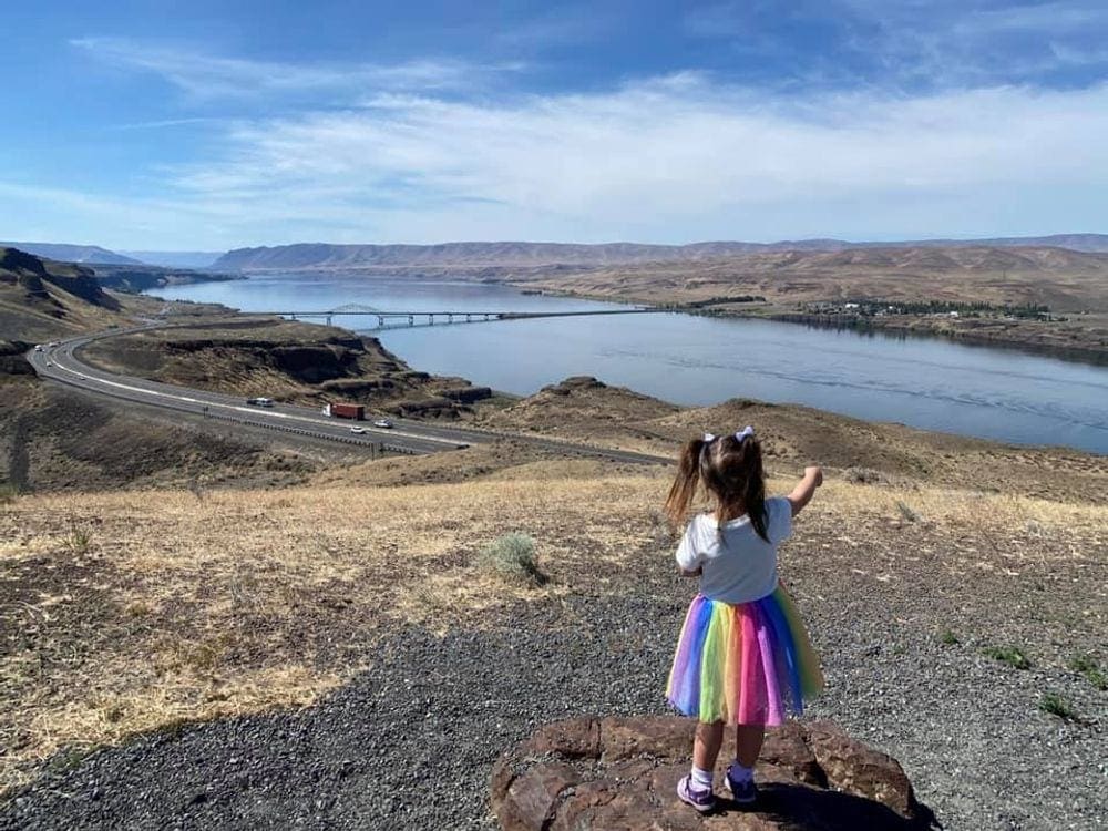 Young girl in rainbow skirt points to river along a road.