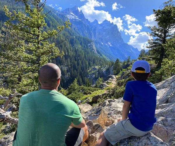 Father and son sit looking out onto the Grand Tetons.
