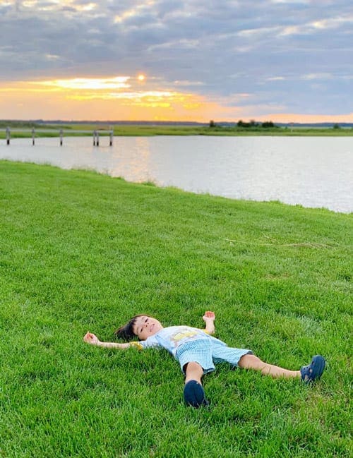 Little boy lying on the grass with water and sunset on the background
