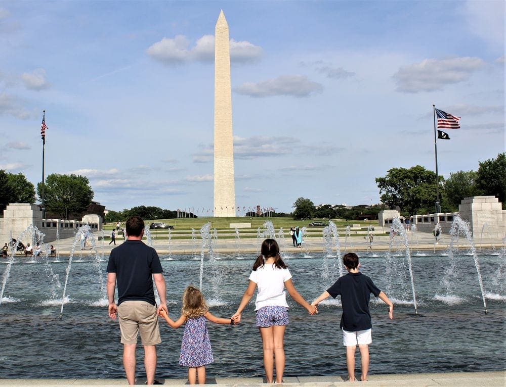 A family of four stands holding hands facing the Washington Monument in the background.