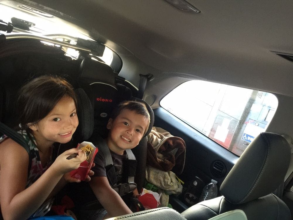 Two kids sit in the back seat of the car enjoying Asian take-out.