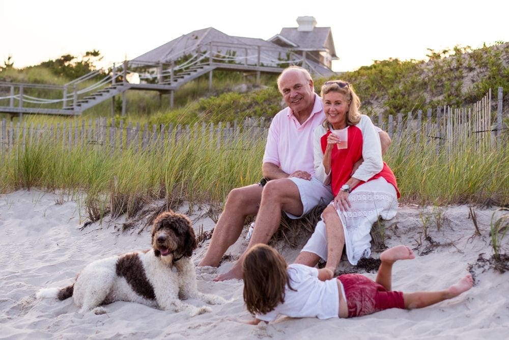 Grandma, grandpa, a young girl, and their dog sit happily on a beach with tall grass and a beach house in the background.