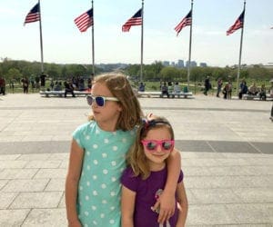 Two girls stand smithing with sunglasses on in front of five American flags in Washington DC.
