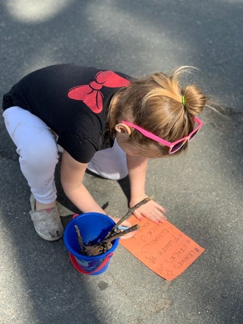 Young girl updates her nature scavenger hunt check list. Near her is her bucket full of collected items, including sticks and leaves.