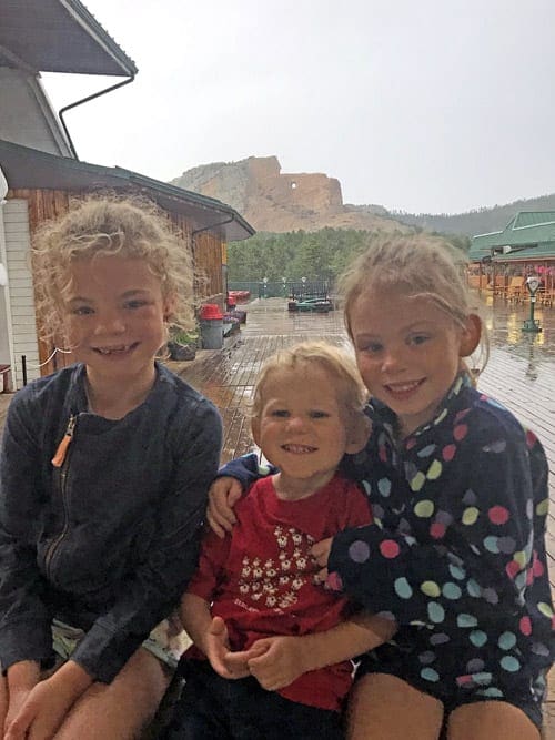 Three young kids sit on a bench with the Crazy Horse Memorial in the distance.