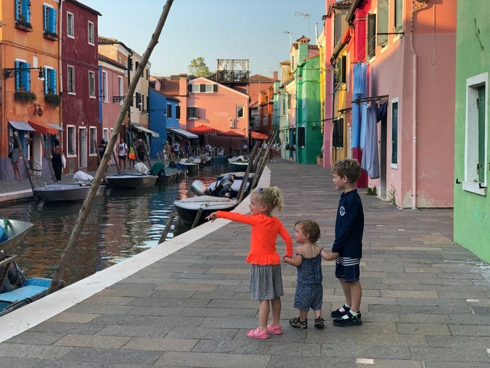 Three small children hold hands along a colorful street in Burano, Italy. One of the girls excitedly points across the canal.