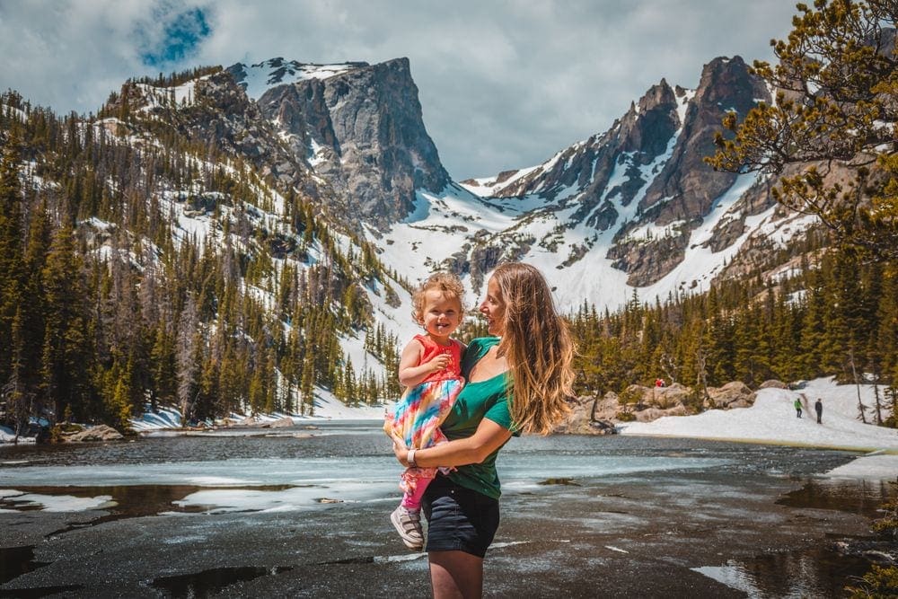 A mom holds her toddler, looking at her fondly, with a ice and snow covered lake and mountains in the background.