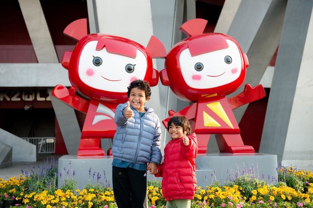 Two young boys stand in front of two red robots in China, both smile at the camera with a thumbs up.