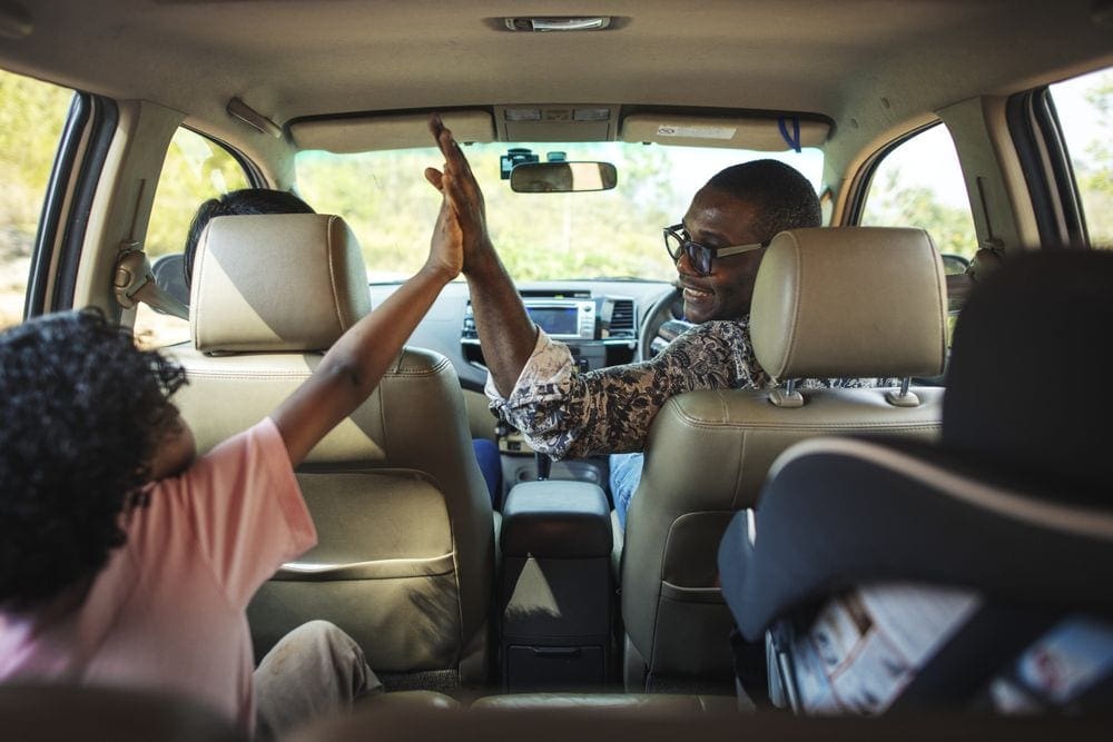 Inside a car, father and child high five while on a road trip, one of the top father-son weekend ideas.