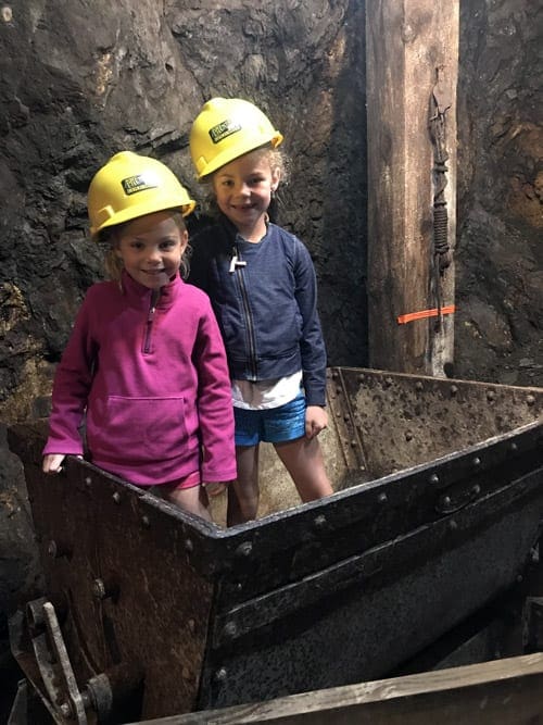 Two young girls stand in a mining cart with yellow hard hats on their heads.