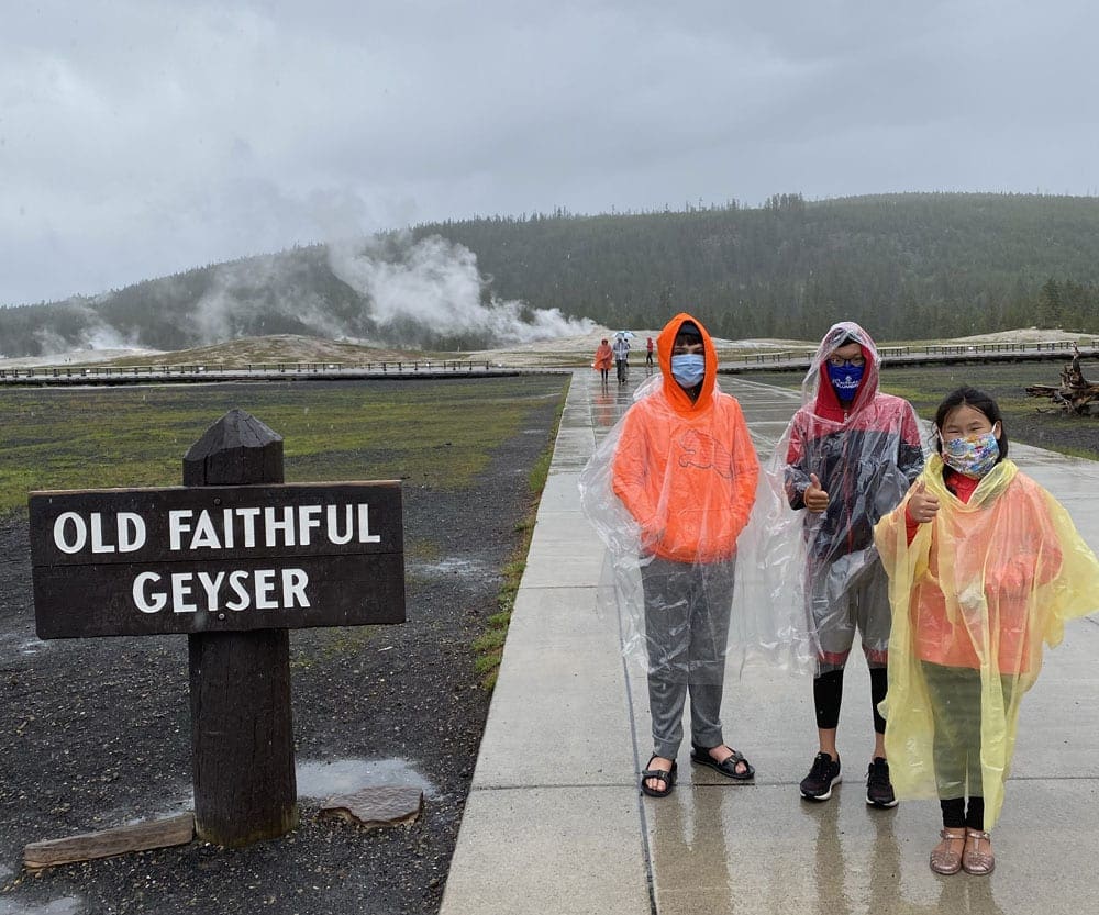 Family of three wearing masks in front of Old Faithful Geysery. Wearing masks is one of the most important Coronavirus Travel Tips For Families.