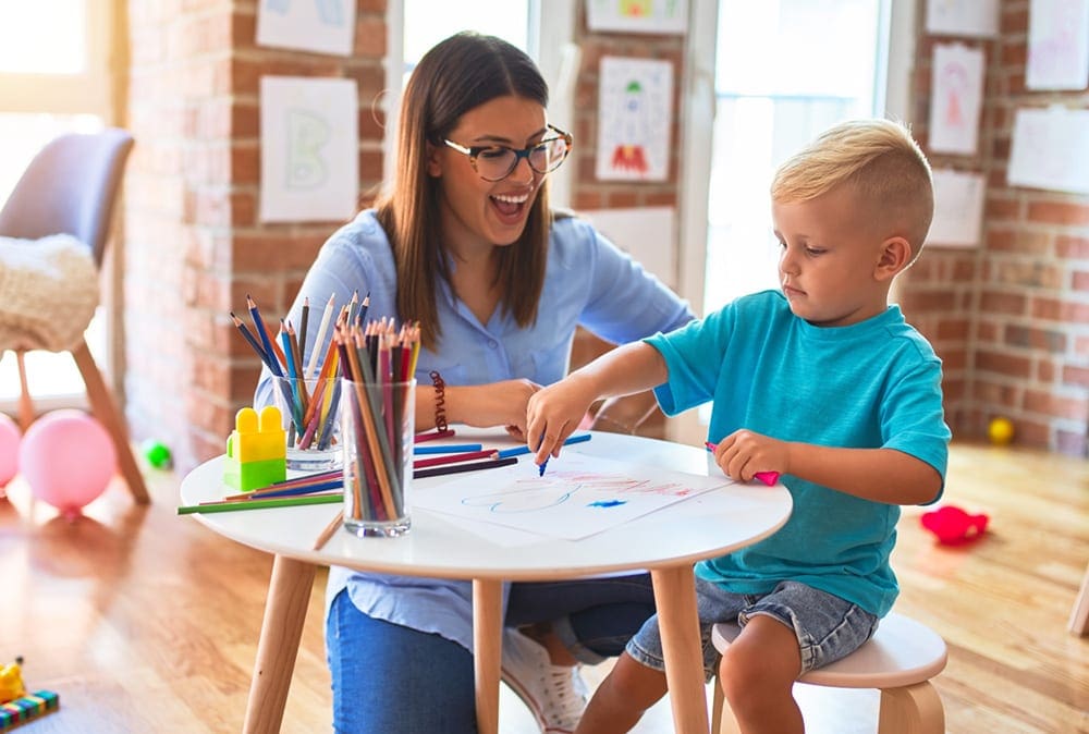 Little boy sitting on a chair, while drawing and talking to an adult. Talking through your plan ahead of time is one of our tips for traveling with children who have allergies.