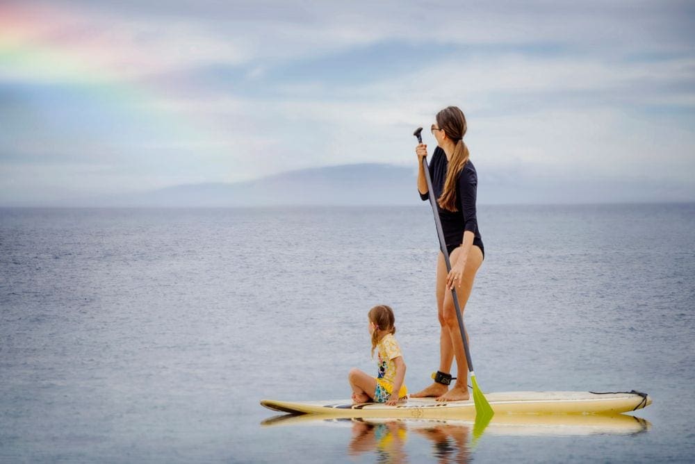 A mother stands on a paddleboard, while her young daughter sits in front of her, off the coast of a Hawaiian beach. A rainbow flows into the water in the background.
