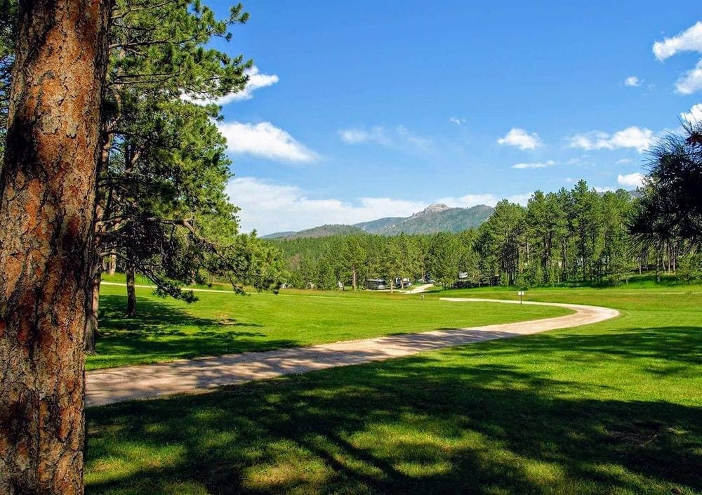 An expansive view of the grounds on Rafter J Bar Ranch Resort, including green grass, a winding path, and mountains in the distance. Rafter J Bar Ranch Resort is a great place to stay while on a Black Hills vacation with kids.
