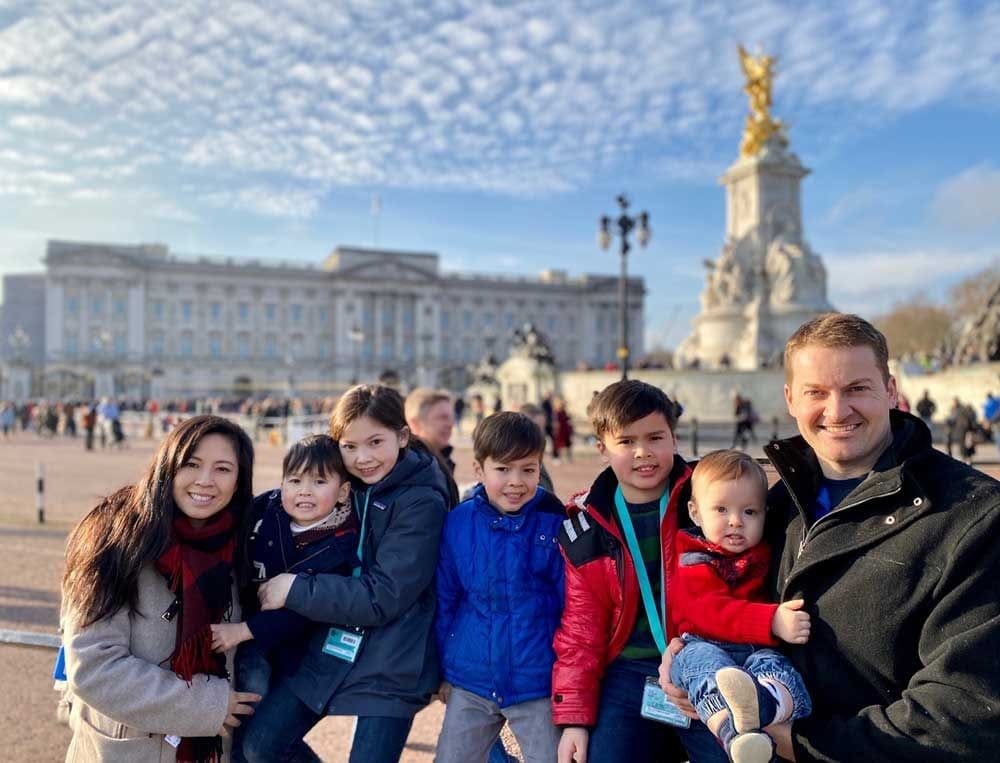 A family of seven sits smiling with Buckingham Palace in the distance, while traveling in London, one of the best places to travel with kids in Europe.