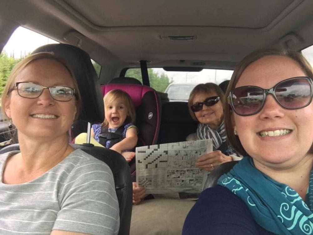 Four generations of women smile while riding in a car, in back a toddler sits in her car seat when traveling.