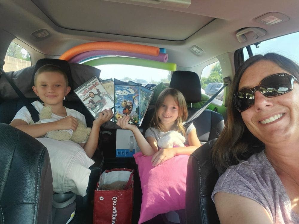A mom and her two kids look excited to embark on a road trip with movies and pool noodles.